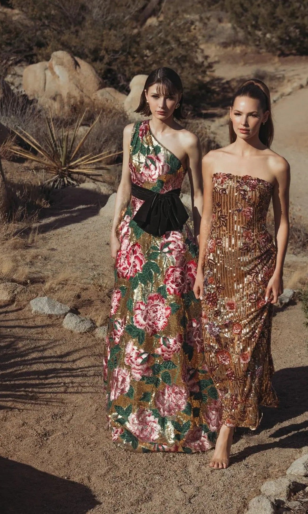 Two women stand barefoot outdoors in the desert, wearing elegant floor-length dresses, including the Mac Duggal 21194 Long Formal Dress with dazzling floral sequin patterns.